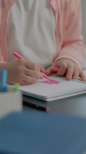 Κάθετη βίντεο: Κοντινό πλάνο του μικρού παιδιού χρησιμοποιώντας πολύχρωμα μολύβια στο σημειωματάριο στο γραφείο. Κοριτσάκι κρατά στυλό για να ζωγραφίσει σκίτσα σε βιβλίο, ενώ κάθεται στο σπίτι μετά την σχολική εργασία. Σχέδιο μαθήτριας - Πλάνα, βίντεο