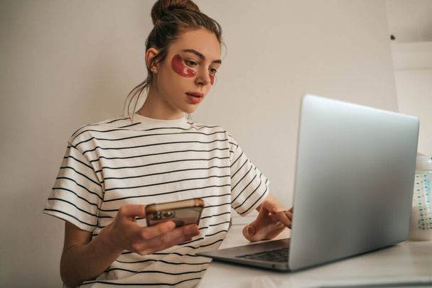 Woman with under-eye patches holding the smartphone in the hand and typing on the laptop - Foto, Bild