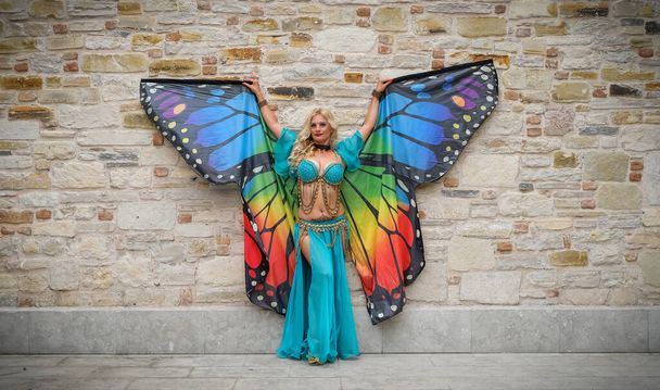 professional belly dancer posing with colorful butterfly wings and authentic dress in front of wall - Photo, Image