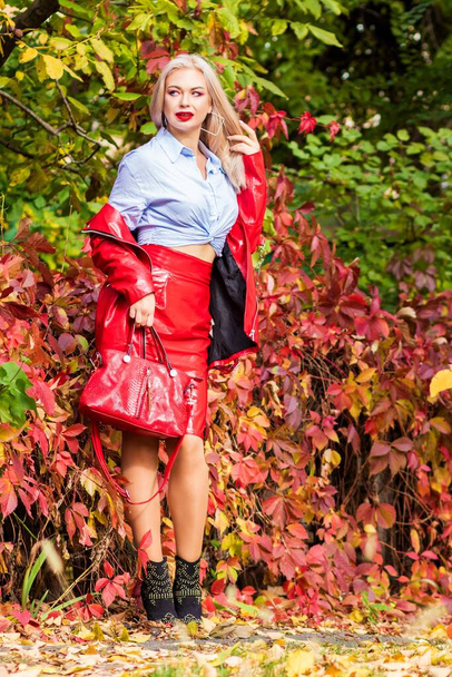 Beautiful stylish woman in blue shirt, red skirt and leather jacket standing and posing in autumn park. Pretty woman with long blonde hairstyle and red lips - Fotoğraf, Görsel