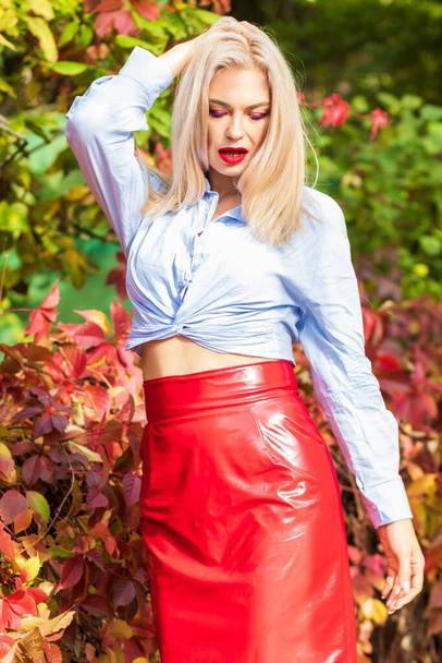 Beautiful stylish woman in blue shirt, red skirt and leather jacket standing and posing in autumn park. Pretty woman with long blonde hairstyle and red lips - Foto, immagini
