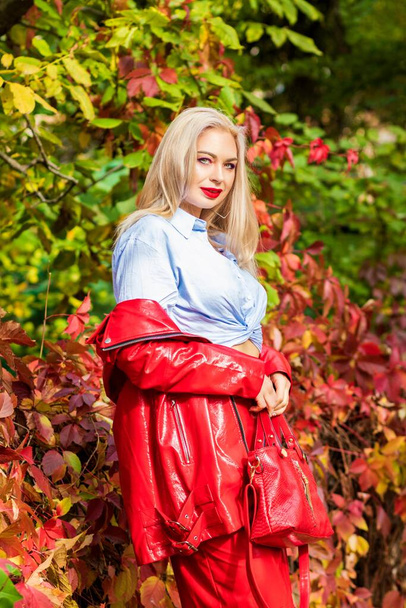Beautiful stylish woman in blue shirt, red skirt and leather jacket standing and posing in autumn park. Pretty woman with long blonde hairstyle and red lips - Фото, изображение