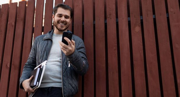 entrepreneur masculin d'âge moyen avec un ordinateur portable dans ses mains et un téléphone portable sur le fond d'une clôture brune. - Photo, image