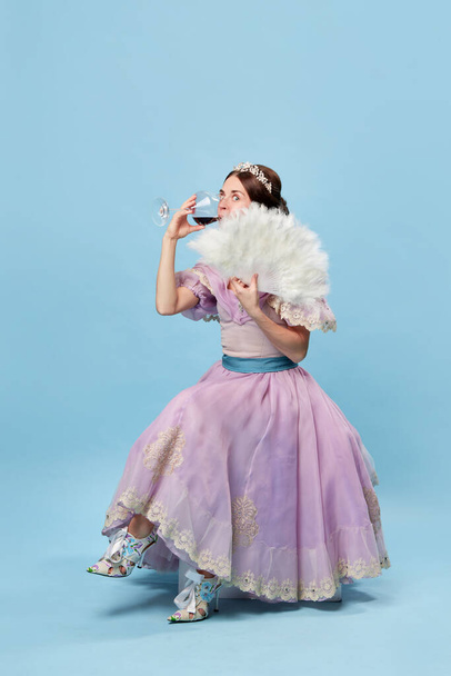 Tasting wine. Cute young girl in lilac color medieval dress as young queen or princess on blue background. Eras comparison, beauty, art, emotions and vintage fashion style. Funny meme emotions - Photo, image