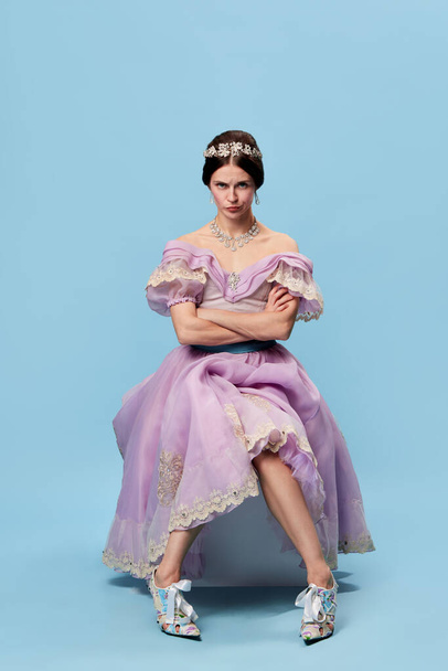 Resentment. Emotional girl in lilac color medieval dress as young queen or princess looking at camera, giving mean look isolated over blue background. Beauty, art, emotions and vintage fashion style - Foto, Imagem