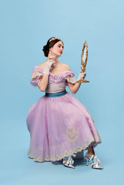 Young and pretty. Beautiful charming woman in lilac color vintage dress as a royal person, princess looking in mirror at herself on blue background. Concept of comparison of eras, renaissance style - Valokuva, kuva