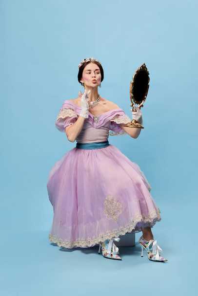 Self loving. Beautiful charming woman in lilac color vintage dress as royal person, princess looking in mirror at herself on blue background. Concept of comparison of eras, modernity and renaissance. - Фото, зображення