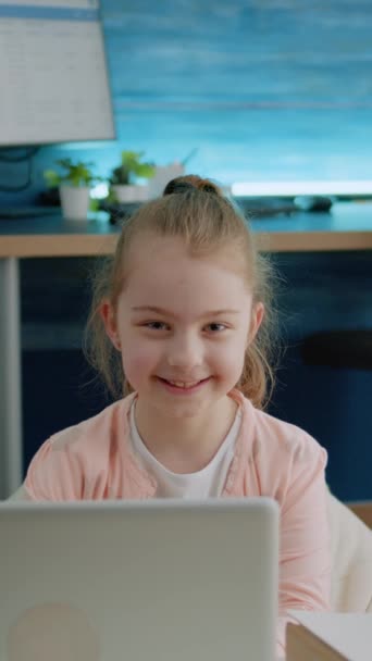 Vidéo verticale : Portrait de jeune fille souriante et tenant un ordinateur portable à la maison pour les devoirs et les cours en ligne. Petit enfant regardant la caméra et se préparant pour les leçons à distance sur l'appareil moderne - Séquence, vidéo