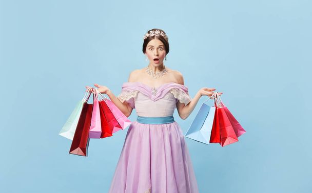Shocked princess. Young woman in image of young queen holding bright shopping packages looking at camera with surprise isolated on blue background. Beauty, black friday, art, sales, vintage style. - Fotografie, Obrázek