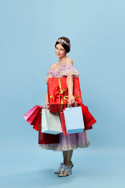 Great shopping day. Beautiful young woman in image of young queen or princess holding colorful shopping bags and festive boxes isolated on blue background. Beauty, black friday, sales, creativity. - Foto, afbeelding
