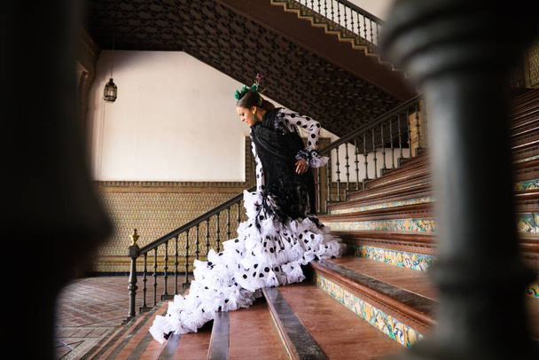 Beautiful teenage woman dancing flamenco with white dress and black polka dots doing flamenco postures on a staircase. She wears a black shawl with fringes. Flamenco cultural heritage of humanity. - Photo, Image