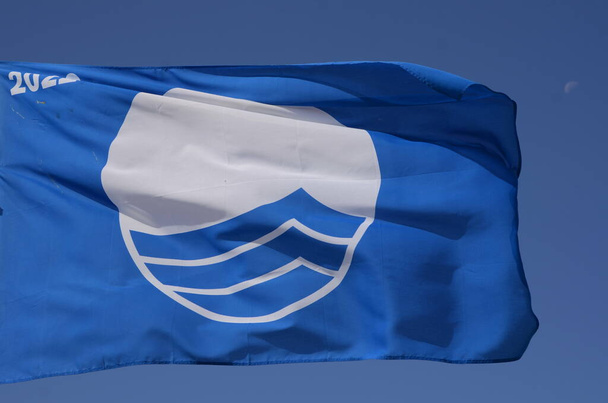空に対する青い旗。世界の海辺にある生態系のビーチのシンボルです。ブルーフラッグは、ビーチやマリーナに対する国際的な賞です。地域の安全・清潔・環境への配慮 - 写真・画像