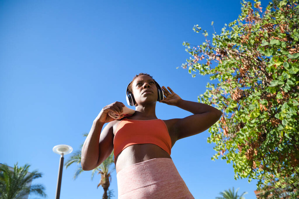 Parkta spor yapan Afro-Amerikalı bir kadın omzuna havlu geçirip beyaz kulaklıklarla müzik dinliyor. O, turuncu bir bluz giydi. Spor ve sağlık kavramı. - Fotoğraf, Görsel