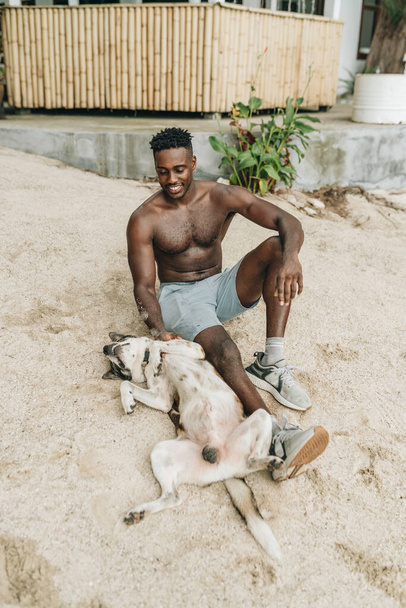 Smiling attractive young black man playing with a dog on the beach. High quality photo - Zdjęcie, obraz