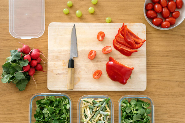 vista superior de pimiento fresco y cuchillo en tablero de madera, tomate en tazón y ensalada de mache en recipiente de vidrio en la mesa en la cocina, preservación de productos alimenticios saludables - Foto, imagen