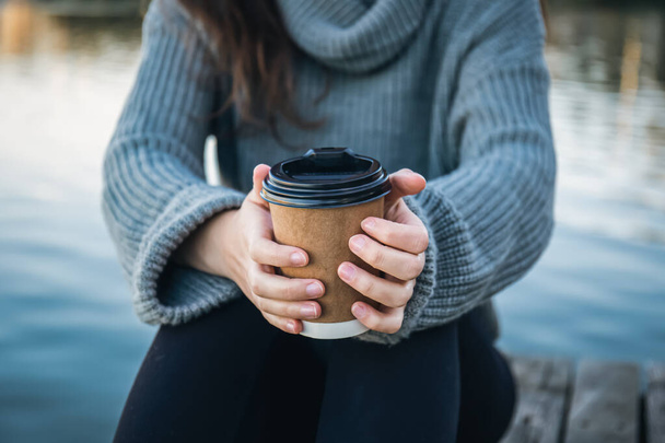 Nahaufnahme, eine Tasse Kaffee in den Händen einer Frau in der Natur am Fluss, ein Getränk zum Mitnehmen in der kalten Jahreszeit. - Foto, Bild