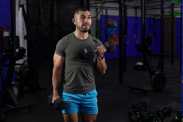 hombre latino con ropa deportiva haciendo ejercicios de bíceps con dos pesas en un gimnasio - Foto, Imagen