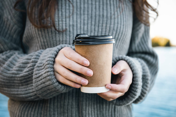 Крупним планом, чашка кави в руках жінки в природі біля річки, напій на винос в холодну пору року
. - Фото, зображення