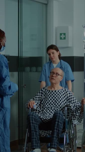 Вертикальне відео: африканський американський лікар бере хворого пацієнта на інвалідному візку від лікарняної палати до операційної. Старий чоловік з IV кап "яним мішком лишає ліжко в блоці з сучасною технологічною медичною технікою. - Кадри, відео