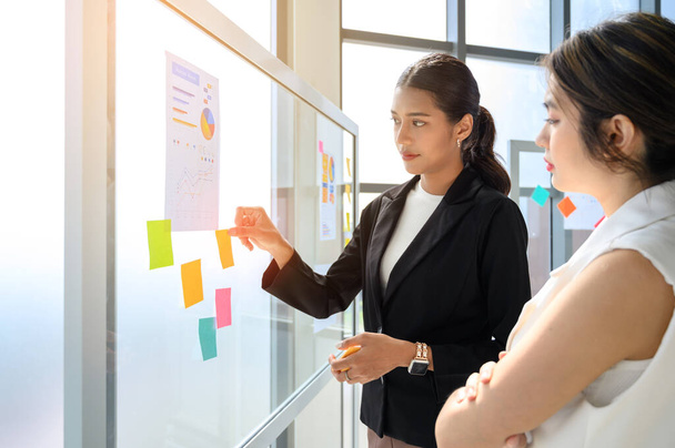 young modern businesswoman in smart casual using sticky notes for strategy ideas while standing behind the glass board in the board room at the office. Business project planning. Hard choice to make. - Photo, image