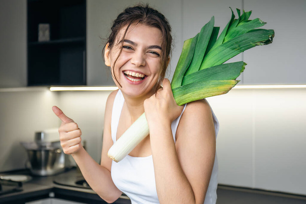 Jovencita alegre en la cocina sosteniendo un puerro en sus manos, el concepto de cocinar y comer sano. - Foto, Imagen