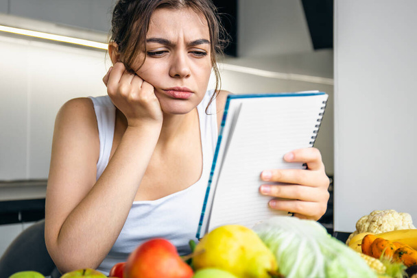 Μια νεαρή γυναίκα στην κουζίνα με ένα σημειωματάριο μεταξύ των λαχανικών, η έννοια της μαγειρικής, δίαιτα και υγιεινή διατροφή. - Φωτογραφία, εικόνα
