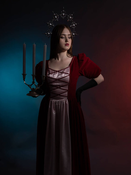 Mythical fantasy queen with a candlestick. Silvery gothic crown on the head. Burgundy vintage artistic dress, black gloves. Medieval style. Portrait in a dark key. Vertical photo. - Фото, зображення