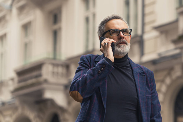 Soustředěný a sebevědomý šedovlasý muž s vousy diskutuje o obchodní dohodě po telefonu, když je na cestě do práce. Kvalitní fotografie - Fotografie, Obrázek