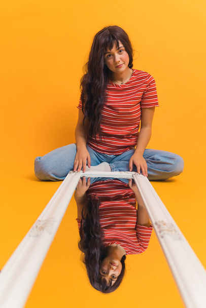 Νεαρό, κομψό κορίτσι με μακριά καστανά μαλλιά κάθεται στο πάτωμα με τον καθρέφτη που βρίσκεται μπροστά της. Γυναικεία αντανάκλαση στον καθρέφτη, απομονωμένο πορτοκαλί φόντο. Υψηλής ποιότητας φωτογραφία - Φωτογραφία, εικόνα