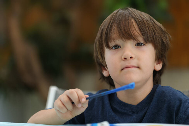 Eine Frontansicht eines niedlichen kleinen Jungen in einem blauen T-Shirt, der draußen sitzt, Pinsel mit blauer Farbe in der Hand hält und in die Kamera blickt - Foto, Bild