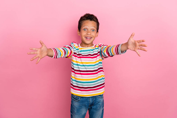 Photo of nice optimistic schoolboy curly hair dressed striped long sleeve holding palms at camera to you isolated on pink color background. - Foto, Imagen