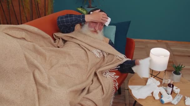 Velho avô doente idoso que sofre de frio ou alergia deitado no sofá em casa. Avô idoso doente com toalha na cabeça sopra espirros toalhetes ranho em guardanapo. Quarentena do vírus da coronária, pandemia - Filmagem, Vídeo