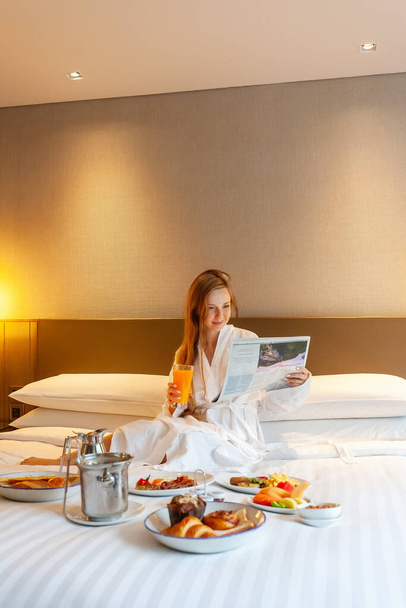 Food in bed serve in luxury hotel. Woman on vacation enjoy breakfast in bed in hotel room, reading newspaper, drinking orange juice, eating food and relaxing on holidays. Room Service - Photo, image