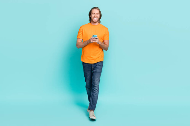 Full length photo of cheerful attractive guy dressed orange t-shirt communicating modern device isolated teal color background. - Zdjęcie, obraz