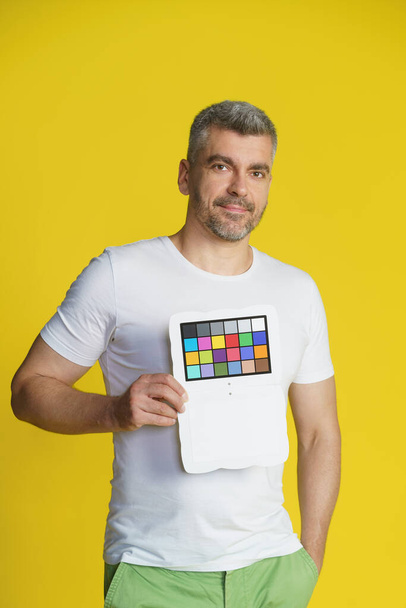 Handsome middle aged model man holding color checker pallet to help photographer with setting up studio wearing white t-shirt isolated on yellow background. Photographer assistant help.  - Foto, Bild