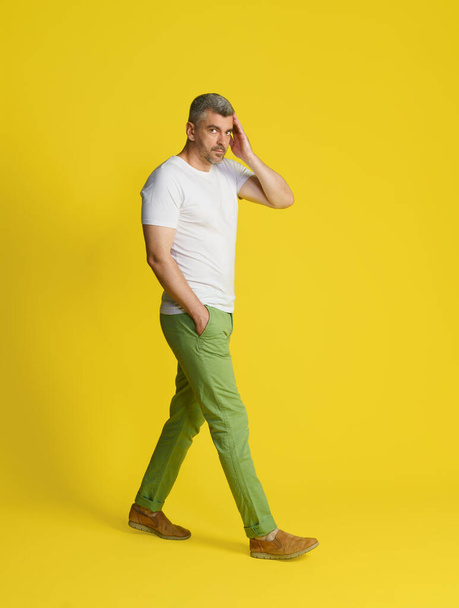 Walking in movement middle aged handsome grey hair man touch face with hand wearing white t-shirt and green jeans isolated on yellow background. Fit, muscular middle aged man. - Fotoğraf, Görsel
