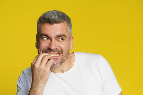 Scared of competitor handsome middle aged man bitting his fingernails. Freelancer in stress of project deadline. Shocked freelancer man eating fingers in white t-shirt isolated on yellow background. - Photo, Image