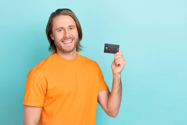 Photo of good mood guy dressed orange t-shirt showing bank card empty space isolated teal color background. - Photo, Image