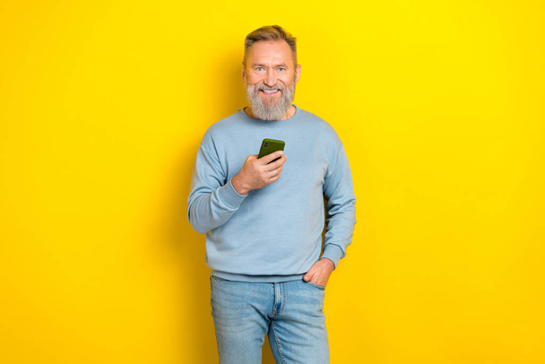 Photo of cute positive retired guy wear blue sweater chatting instagram twitter facebook telegram gadget isolated yellow color background. - Foto, imagen