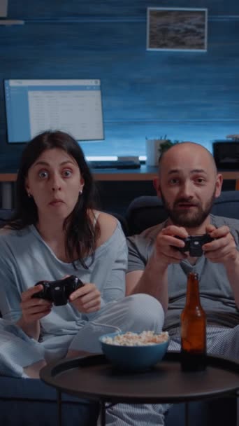 Κάθετο βίντεο: Κατεστραμμένο ενθουσιασμένο ζευγάρι που κερδίζει βιντεοπαιχνίδια, αγκαλιασμένοι καθισμένοι στον καναπέ παίζοντας, η ευτυχία ουρλιάζει απολαμβάνοντας τον online ανταγωνισμό. Ανταγωνιστικοί παίκτες που χρησιμοποιούν joysticks playstation - Πλάνα, βίντεο