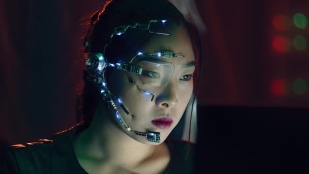 Una chica Cyberpunk mira la pantalla de la computadora. Usando gafas futuristas de un solo ojo con micrófono. Deja de mirar a la computadora para mirar a la cámara. Fondos cibernéticos y de ciencia ficción. - Imágenes, Vídeo