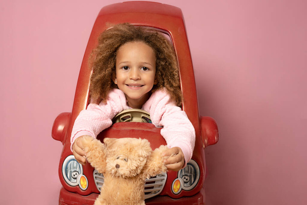 可愛いです小さな子供の女の子運転子供の電動おもちゃ車保持Aテディベア上のピンクの背景 - 写真・画像