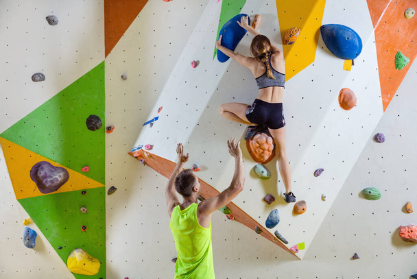 Rock climbers in climbing gym. Young woman climbing bouldering problem (route), male instructor securing her. - Photo, Image