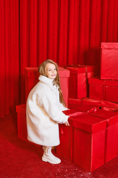Cute little girl in white coat by the huge red christmas presents boxes on red background. Holidays, shopping, fun, fashion - Fotoğraf, Görsel