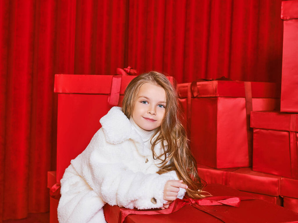 Симпатична маленька дівчинка в білому пальто від величезного червоного Різдва представляє коробки на червоному тлі. Свята, покупки, веселощі, мода
 - Фото, зображення