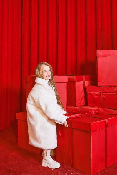 Cute little girl in white coat by the huge red christmas presents boxes on red background. Holidays, shopping, fun, fashion - Foto, imagen