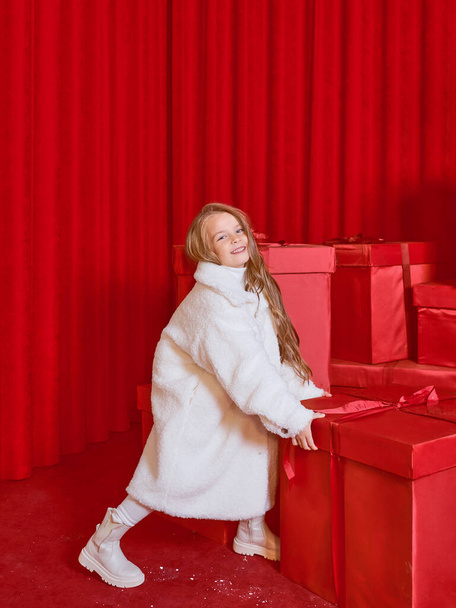 Menina bonito no casaco branco pelo enorme Natal vermelho apresenta caixas no fundo vermelho. Férias, compras, diversão, moda - Foto, Imagem