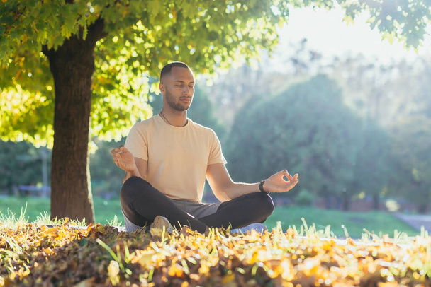 Ein junger Mann meditiert im Park bei Sonnenuntergang, ein Athlet in Lotusposition sitzt auf einer Sportmatte unter den Herbstbäumen. - Foto, Bild