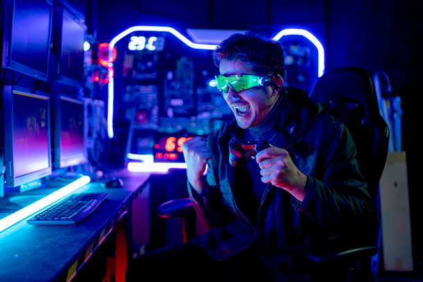 Kaukázusi Gamer férfi hitech ruhák és szemüveg mutatják fellépés izgatott és boldog játék közben számítógépes játék a szobában sötét és többszínű fény, hogy össze hűvös érzelem. - Fotó, kép
