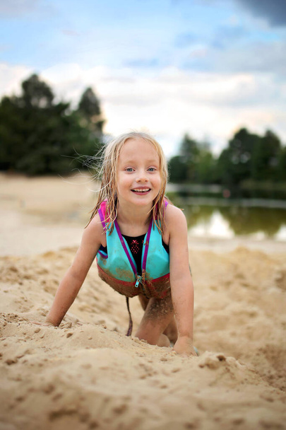 Ein fröhliches kleines Mädchen mit Schwimmweste spielt draußen im Sand am Strand am Badesee. - Foto, Bild